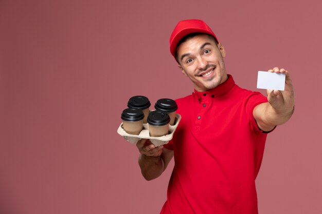 Männlicher Kurier der Vorderansicht in der roten Uniform, die Lieferkaffeetassen mit der weißen Karte hält, die auf rosa Wanddienstlieferungsjobarbeiteruniform lächelt