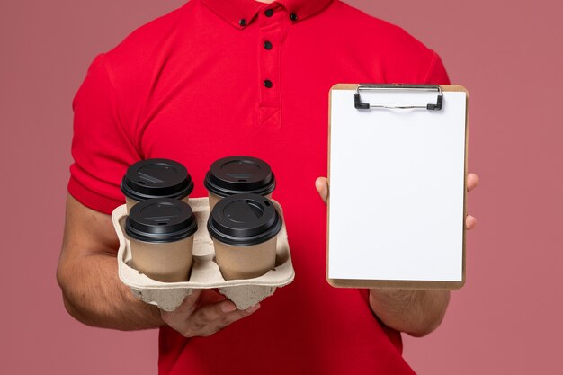 Männlicher Kurier der Vorderansicht in der roten Uniform, die Lieferkaffeetassen mit Notizblock an der rosa Wand hält