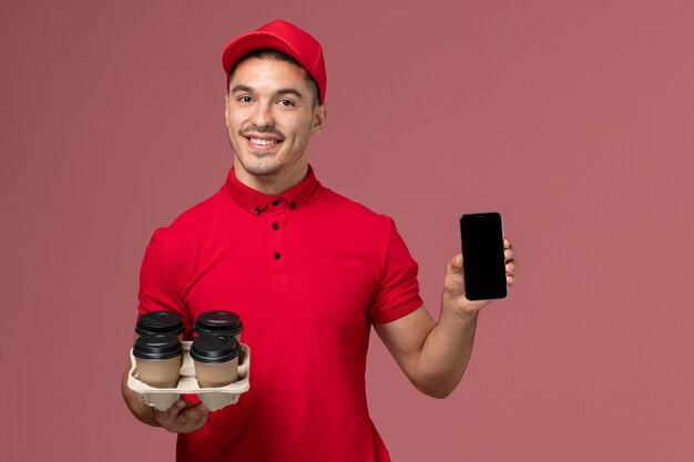 Männlicher Kurier der Vorderansicht in der roten Uniform lächelnd und hält Lieferkaffeetassen mit Telefon auf rosa Wand