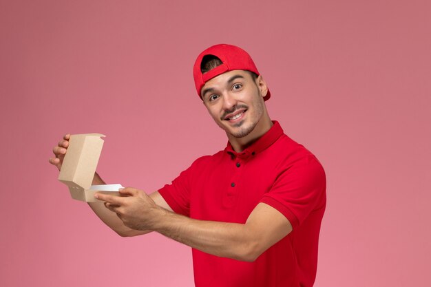 Männlicher Kurier der Vorderansicht in der roten Uniform und im Umhang, der kleines Lieferpaket auf dem rosa Schreibtisch hält.