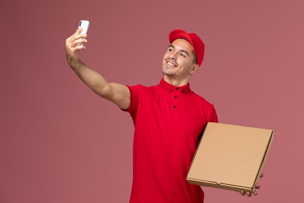 Männlicher Kurier der Vorderansicht in der roten Uniform und im Umhang, der Nahrungsmittelbox hält, die Foto mit ihm auf männlicher Lieferuniform des rosa Wanddienstauftrags nimmt