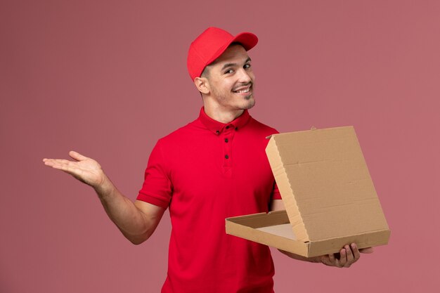 Männlicher Kurier der Vorderansicht in der roten Uniform und im Umhang, der Nahrungsmittelbox mit Lächeln auf hellrosa Wandarbeiter hält