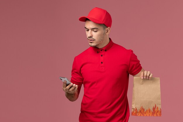 Männlicher Kurier der Vorderansicht in der roten Uniform und im Umhang, der Nahrungsmittelpaket hält und sein Telefon an der rosa Wand benutzt