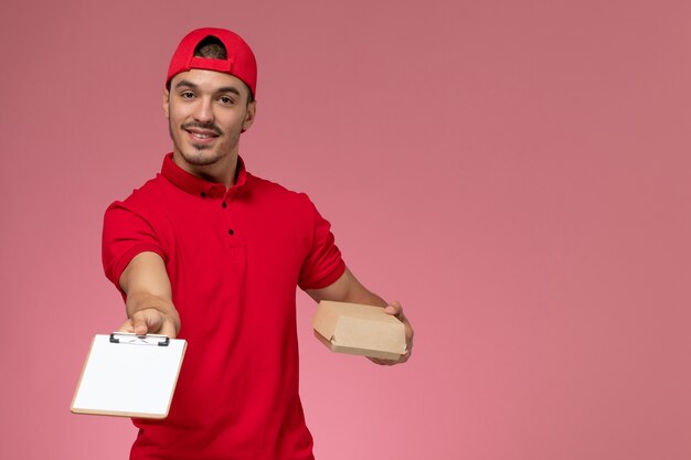 Männlicher Kurier der Vorderansicht in der roten Uniform und im Umhang, die kleines Lieferpaket mit Notizblock lächelnd auf dem rosa Hintergrund halten.