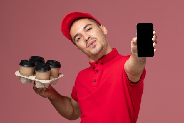Männlicher Kurier der Vorderansicht in der roten Uniform und im Umhang, die Lieferkaffeetassen mit Telefon auf hellrosa Wand halten