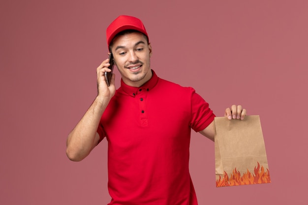 Männlicher Kurier der Vorderansicht in der roten Uniform und im Umhang, die Nahrungsmittelpaket halten und am Telefon an der rosa Wand sprechen