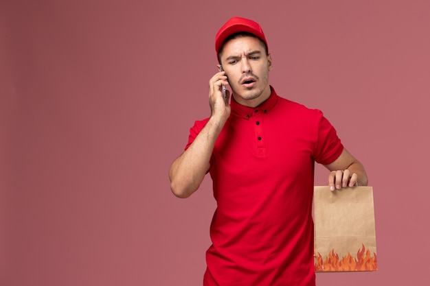 Männlicher Kurier der Vorderansicht in der roten Uniform und im Umhang, die Nahrungsmittelpaket halten und am Telefon an der rosa Wand sprechen