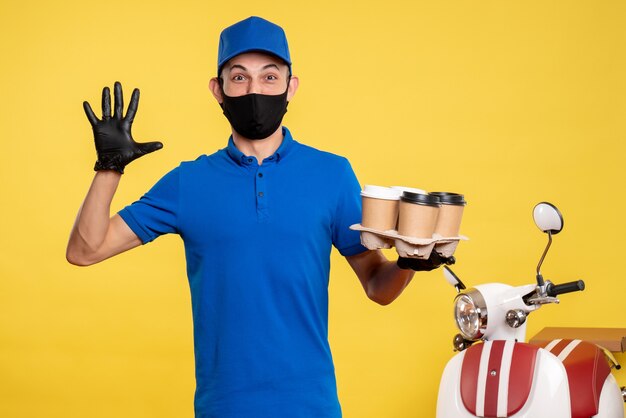 Männlicher Kurier der Vorderansicht in der schwarzen Maske, die Kaffee auf gelbem Job-Covid-Pandemie-Zustelldienst hält