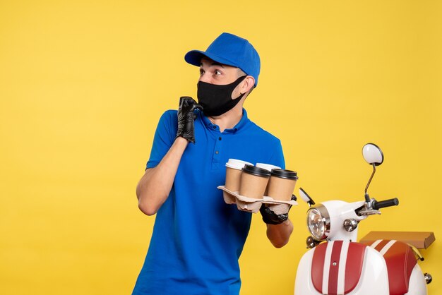 Männlicher Kurier der Vorderansicht in der schwarzen Maske, die Kaffee auf gelbem Job kovidpandemischer einheitlicher Arbeitsdienst hält