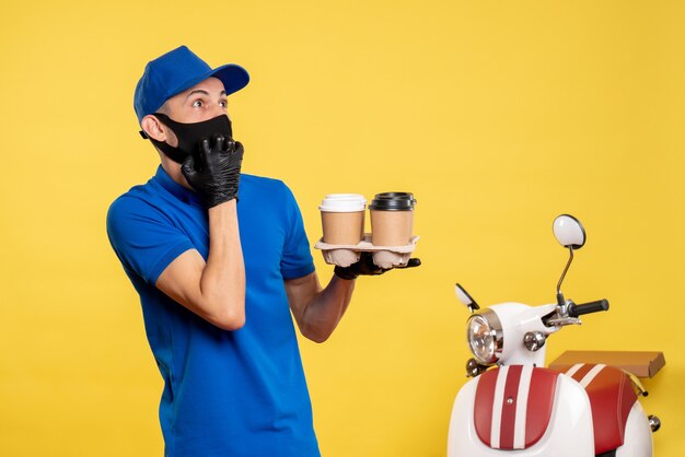 Männlicher kurier der vorderansicht in der schwarzen maske, die kaffee auf gelber arbeit lieferung job covid pandemie service uniform hält