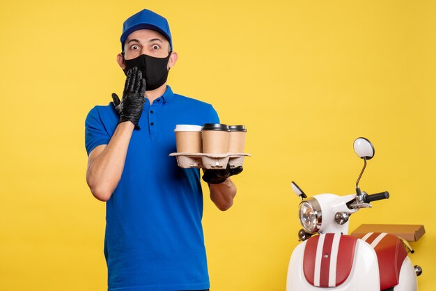 Männlicher Kurier der Vorderansicht in der schwarzen Maske, die Kaffee auf gelber Arbeit Lieferung Job Covid Pandemie Uniform hält