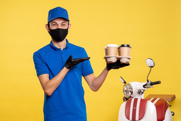 Männlicher Kurier der Vorderansicht in der schwarzen Maske, die Kaffee auf gelber kovidpandemischer Arbeitsuniform des Lieferauftrags hält