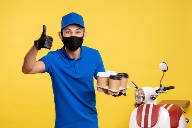 Männlicher Kurier der Vorderansicht in der schwarzen Maske, die Kaffee auf gelber Lieferung Job Covid-Work Service Uniform hält