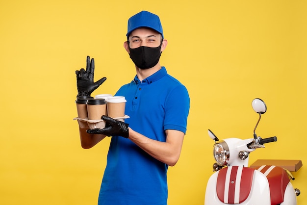 Männlicher Kurier der Vorderansicht in der schwarzen Maske, die Kaffee auf Uniformarbeit der gelben Job-Covid-Pandemie-Dienstlieferung hält