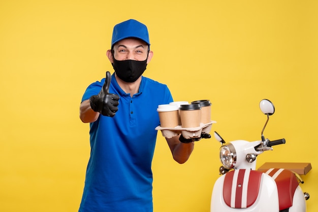 Männlicher Kurier der Vorderansicht in der schwarzen Maske, die Kaffee hält und auf gelber Arbeit Lieferung Job Covid Pandemie Service Uniform lachen