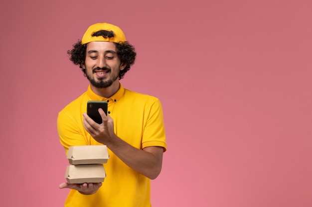 Männlicher Kurier der Vorderansicht in gelbem Uniformumhang mit Lebensmittelpaketen auf seinen Händen, die am Telefon auf hellrosa Hintergrund sprechen.