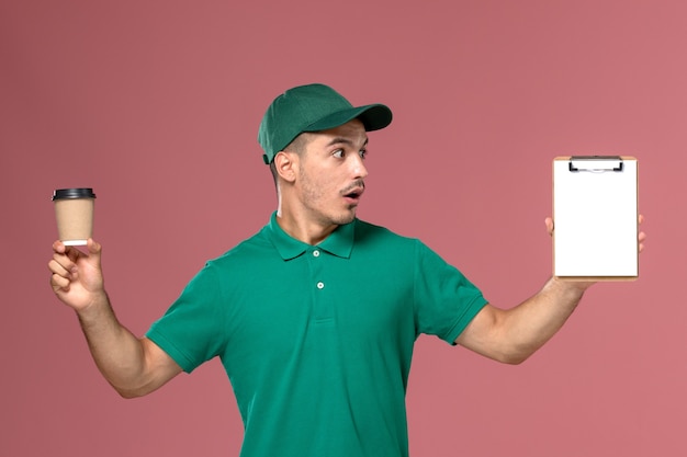 Männlicher Kurier der Vorderansicht in grüner Uniform, die Lieferung Kaffeetasse und Notizblock auf rosa Hintergrund hält