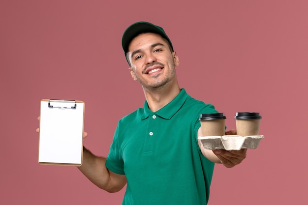 Männlicher Kurier der Vorderansicht in grüner Uniform mit braunen Kaffeetassen und Notizblock auf dem hellrosa Schreibtisch