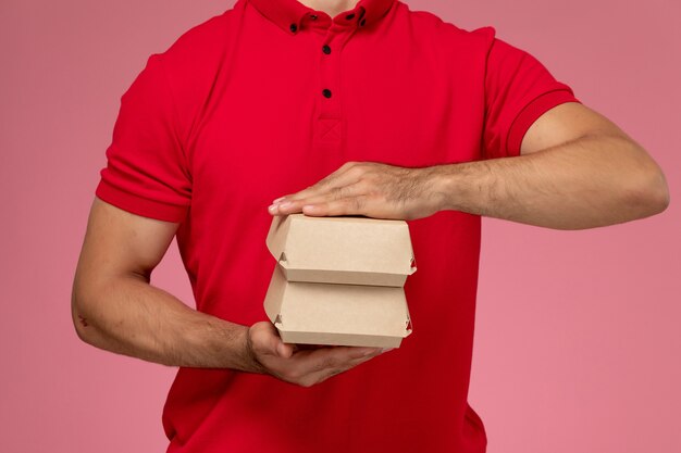 Männlicher Kurier der Vorderansicht in roter Uniform und Kappe, die kleines Lieferpaket an der rosa Wand halten