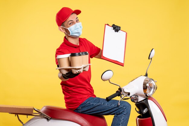Männlicher Kurier der Vorderansicht in roter Uniform und Maske mit Kaffee auf gelbem Jobpandemie-Lieferfarbkovid-Arbeitsnahrungsmittel