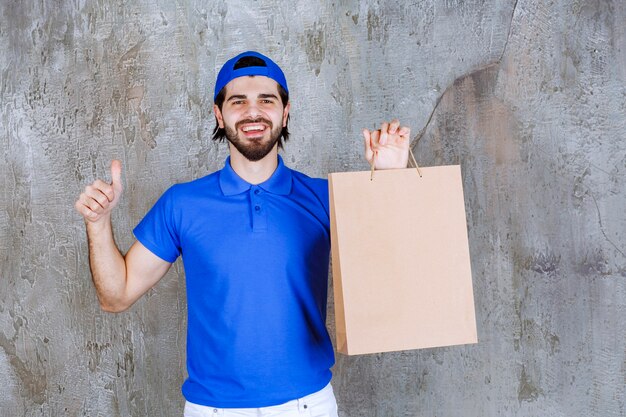 Männlicher Kurier in blauer Uniform, der eine Einkaufstüte aus Pappe hält und ein positives Handzeichen zeigt