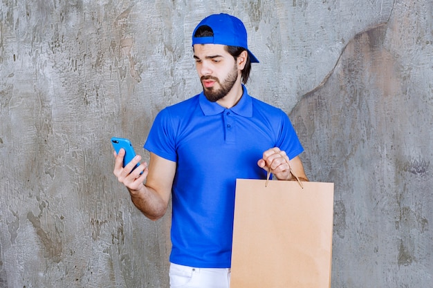 Männlicher Kurier in blauer Uniform, der eine Einkaufstüte aus Pappe hält und mit dem Telefon spricht.