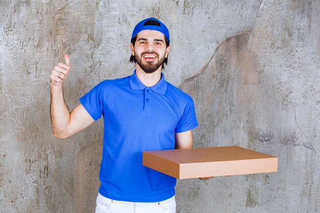 Männlicher Kurier in blauer Uniform, der eine Pappschachtel zum Mitnehmen trägt und ein positives Handzeichen zeigt.