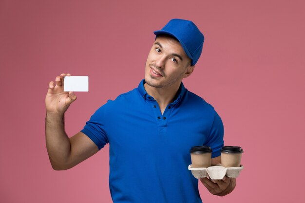 männlicher Kurier in blauer Uniform, die Kaffeetassen und Karte auf rosa, Arbeiteruniform-Dienstlieferung hält