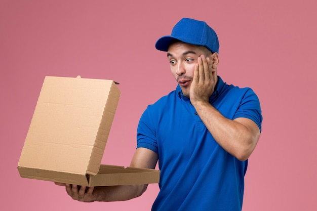 männlicher Kurier in blauer Uniform, die Lebensmittelbox hält, die es auf rosa, einheitlicher Dienstauftragslieferung schockiert öffnet