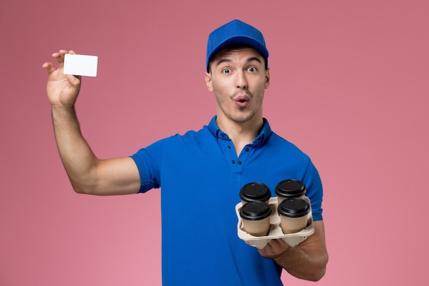 männlicher Kurier in blauer Uniform, die Lieferkaffeetassen und Karte auf rosa, einheitlicher Jobarbeiterdienstlieferung hält