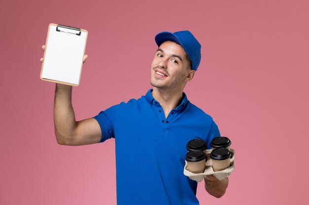 männlicher Kurier in blauer Uniform mit Lieferkaffeetassen und Notizblock auf rosa, einheitlicher Servicebereitstellung