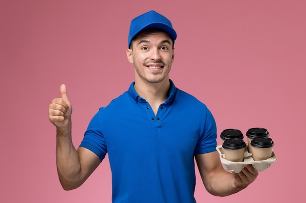 männlicher Kurier in der blauen Uniform, die braune Kaffeetassen hält und auf rosa, Arbeiteruniform-Dienstlieferung lächelt