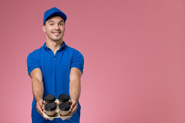 männlicher Kurier in der blauen Uniform, die braune Kaffeetassen hält und auf rosa, Arbeiteruniform-Dienstlieferung lächelt