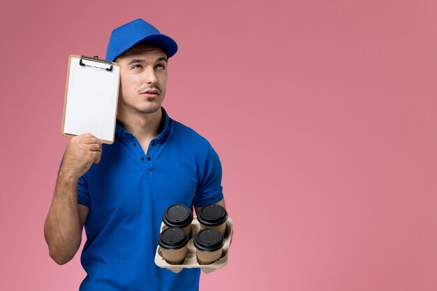 männlicher Kurier in der blauen Uniform, die braune Kaffeetassen mit Notizblock hält, der auf rosa, einheitliche Servicebereitstellung denkt