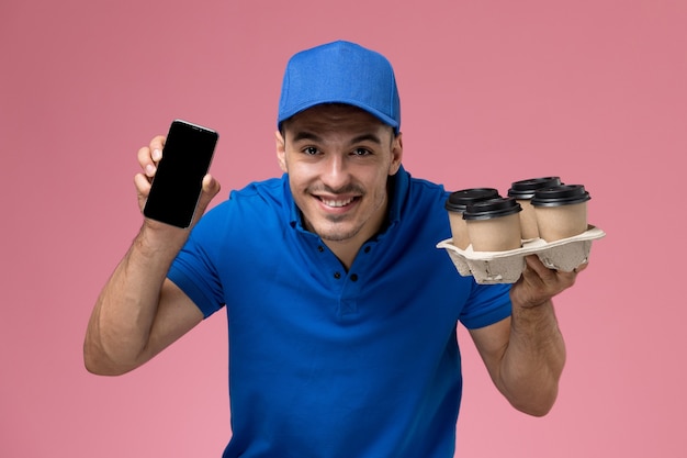 männlicher Kurier in der blauen Uniform, die braune Kaffeetassen mit Telefon auf rosa, Arbeiteruniform-Dienstlieferung hält
