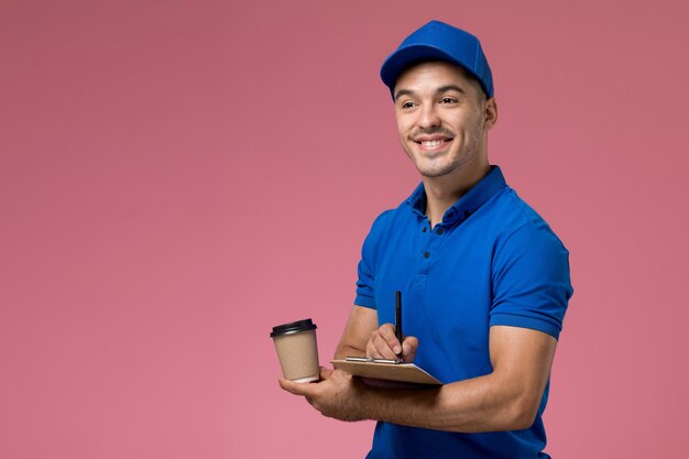 männlicher Kurier in der blauen Uniform, die Kaffeetasse und Notizblock mit Lächeln auf rosa, Arbeiteruniform-Dienstlieferung hält