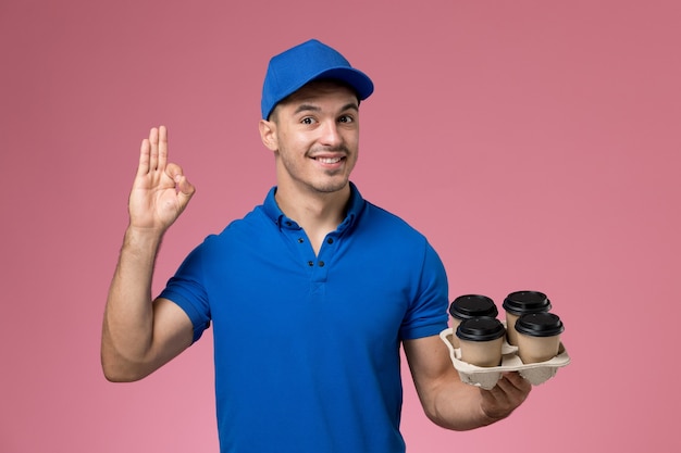 männlicher Kurier in der blauen Uniform, die Kaffeetassen hält und auf rosa, Arbeiteruniform-Dienstlieferung aufwirft