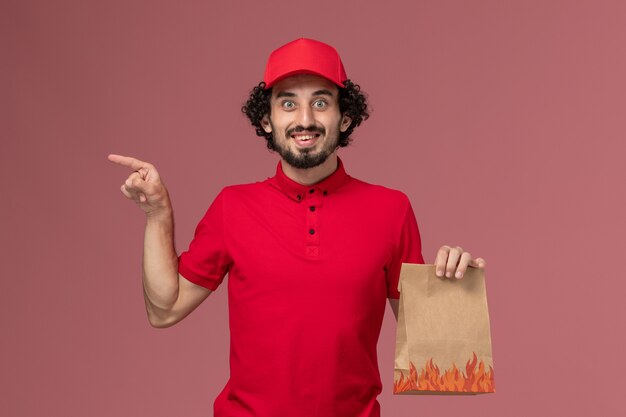 Männlicher Kurierlieferant der Vorderansicht im roten Hemd und im Umhang, der Lebensmittelpaket auf rosa Wanddienstlieferungsjobarbeitsarbeitsangestellter hält