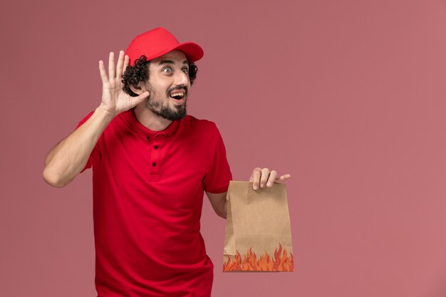 Männlicher Kurierlieferant der Vorderansicht im roten Hemd und im Umhang, der Papiernahrungsmittelpaket hält, das versucht, auf rosa Wanddienstlieferungsmitarbeiter zu hören