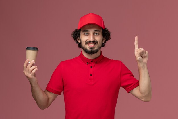 Männlicher Kurierlieferant der Vorderansicht im roten Hemd und im Umhang, die braune Kaffeetasse auf hellrosa Wanddienstuniformlieferungsangestellter männlicher Arbeiter halten