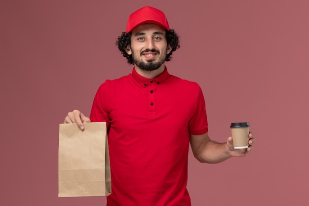 Männlicher Kurierlieferant der Vorderansicht im roten Hemd und im Umhang, die braune Kaffeetasse und Lebensmittelpaket auf hellrosa Wandservice-Zustellungsmitarbeiter halten