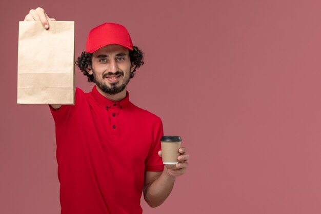 Männlicher Kurierlieferant der Vorderansicht im roten Hemd und im Umhang, die braune Kaffeetasse und Nahrungsmittelpaket auf hellrosa Wanddienstlieferungsarbeitsmitarbeiterjob halten