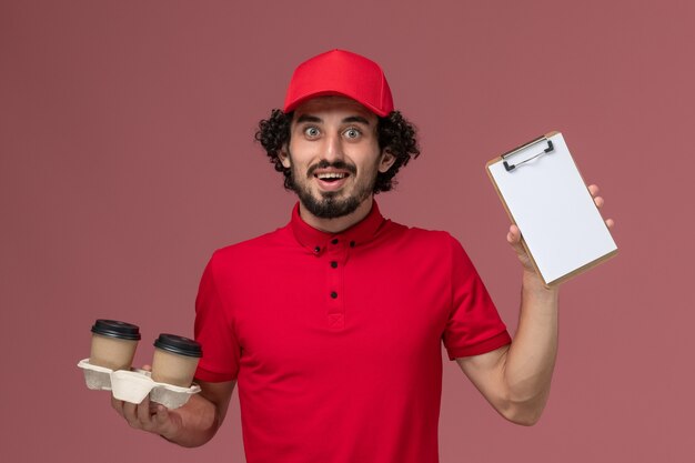 Männlicher Kurierlieferant der Vorderansicht im roten Hemd und im Umhang, die braune Lieferkaffeetassen und Notizblock auf der hellrosa Wandarbeitslieferungsmitarbeiterjobarbeit halten