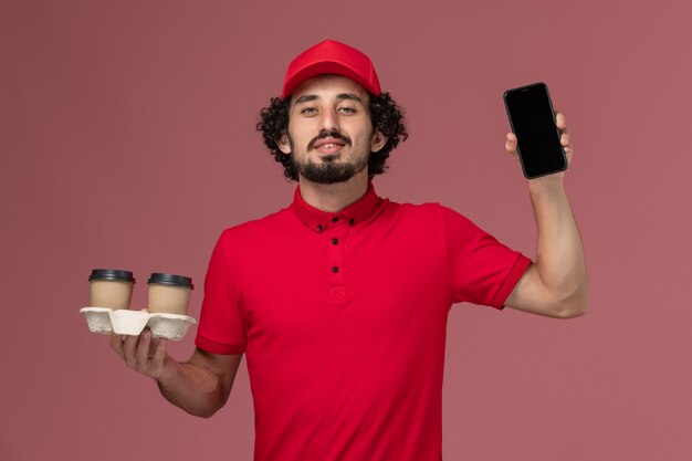 Männlicher Kurierlieferant der Vorderansicht im roten Hemd und im Umhang, die braune Lieferkaffeetassen und Telefon auf dem hellrosa Wanddienstlieferungsmitarbeiterjob halten