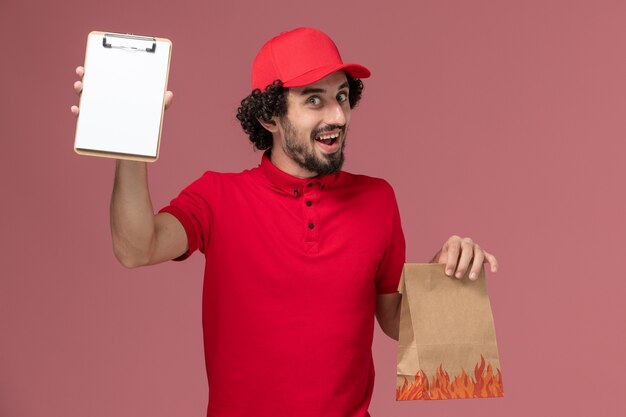 Männlicher Kurierlieferant der Vorderansicht im roten Hemd und im Umhang, die Lebensmittelpaket und Notizblock auf dem rosa Wanddienstlieferungsmitarbeiterarbeitsmann halten