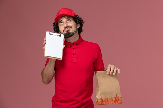 Männlicher Kurierlieferant der Vorderansicht im roten Hemd und im Umhang, die Lebensmittelpaket und Notizblock halten, die gerade auf den rosa Wanddienstlieferungsmitarbeiter denken