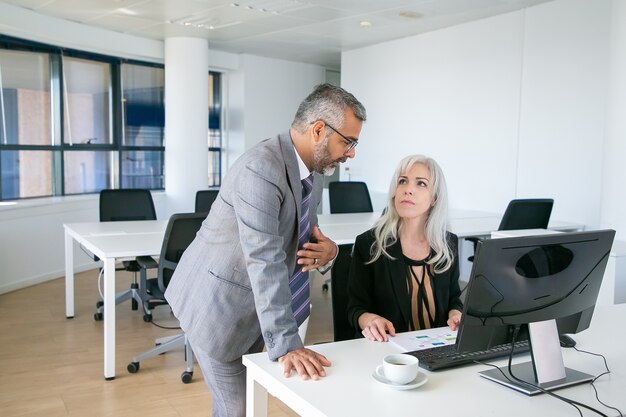 Männlicher Manager erklärt seinen Bericht der Chefin. Kollegen sitzen und stehen am Arbeitsplatz mit PC und diskutieren Diagramm. Geschäftskommunikationskonzept