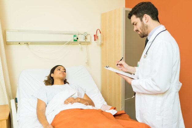 Männlicher Mitarbeiter des Gesundheitswesens, der Berichte über die Zwischenablage erstellt, während er neben einer Patientin steht, die im Krankenhaus im Bett liegt