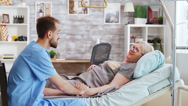 Männlicher Pfleger, der mit einer kranken alten Dame im Krankenhausbett spricht, nimmt die Hand der Frau