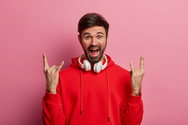 Männlicher rocker genießt positive stimmung, hört rock'n'roll, coole musik im club, verwendet moderne stereokopfhörer, trägt einen roten hoodie, posiert an einer rosigen pastellwand und zeigt eine horngeste. körpersprache